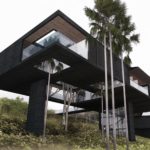 Exterior perspective rendering of a modern hillside black like tree house; day mode from down angular view.