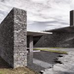 Exterior photography of stone walls and path via a waterfront to the entrance of a minimalist stone mosque; day mode angular view.