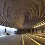 Interior photography of minimalist concrete mosque with stone walls, gradual scaled ceiling with linear skylight, and linear long steps with LED lights.