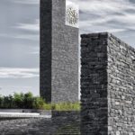 Exterior photography of stone walls and a minaret (vertical minimalist stone element) of a minimalist stone mosque; day mode angular view.