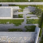Exterior photography of hillside partially underground minimalist stone mosque with green roof; day mode from above view.