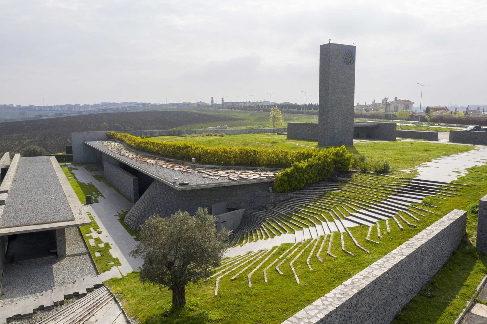 Exterior photography of hillside partially underground minimalist stone mosque with green roof; day mode angular view.