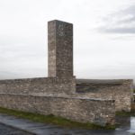 Exterior photography of stone walls and a minaret (vertical minimalist stone element) of a minimalist stone mosque; day mode angular view.