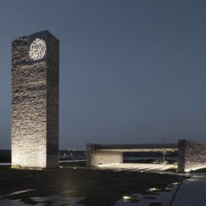 Exterior photography of stone walls and a minaret (vertical minimalist stone element) of a minimalist stone mosque; night mode angular view.