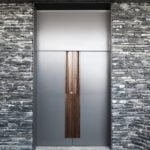 Exterior photography of a metallic frontal door in between stone walls; day mode frontal view.