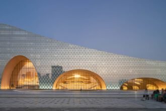 A modern architectural building with a unique design featuring large, curved archways and a facade covered in a geometric pattern of triangular shapes. The building emits a warm glow from within through its glass windows, creating a sleek and reflective contrast against the evening sky. A paved plaza is visible in the foreground, with a few people sitting and walking, which adds a sense of scale to the structure.