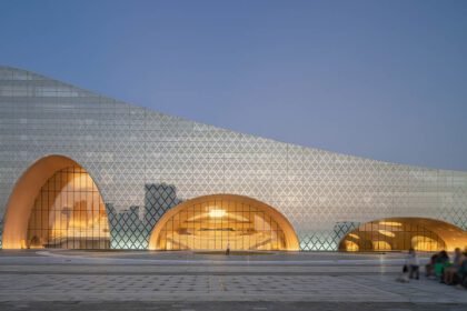 A modern architectural building with a unique design featuring large, curved archways and a facade covered in a geometric pattern of triangular shapes. The building emits a warm glow from within through its glass windows, creating a sleek and reflective contrast against the evening sky. A paved plaza is visible in the foreground, with a few people sitting and walking, which adds a sense of scale to the structure.