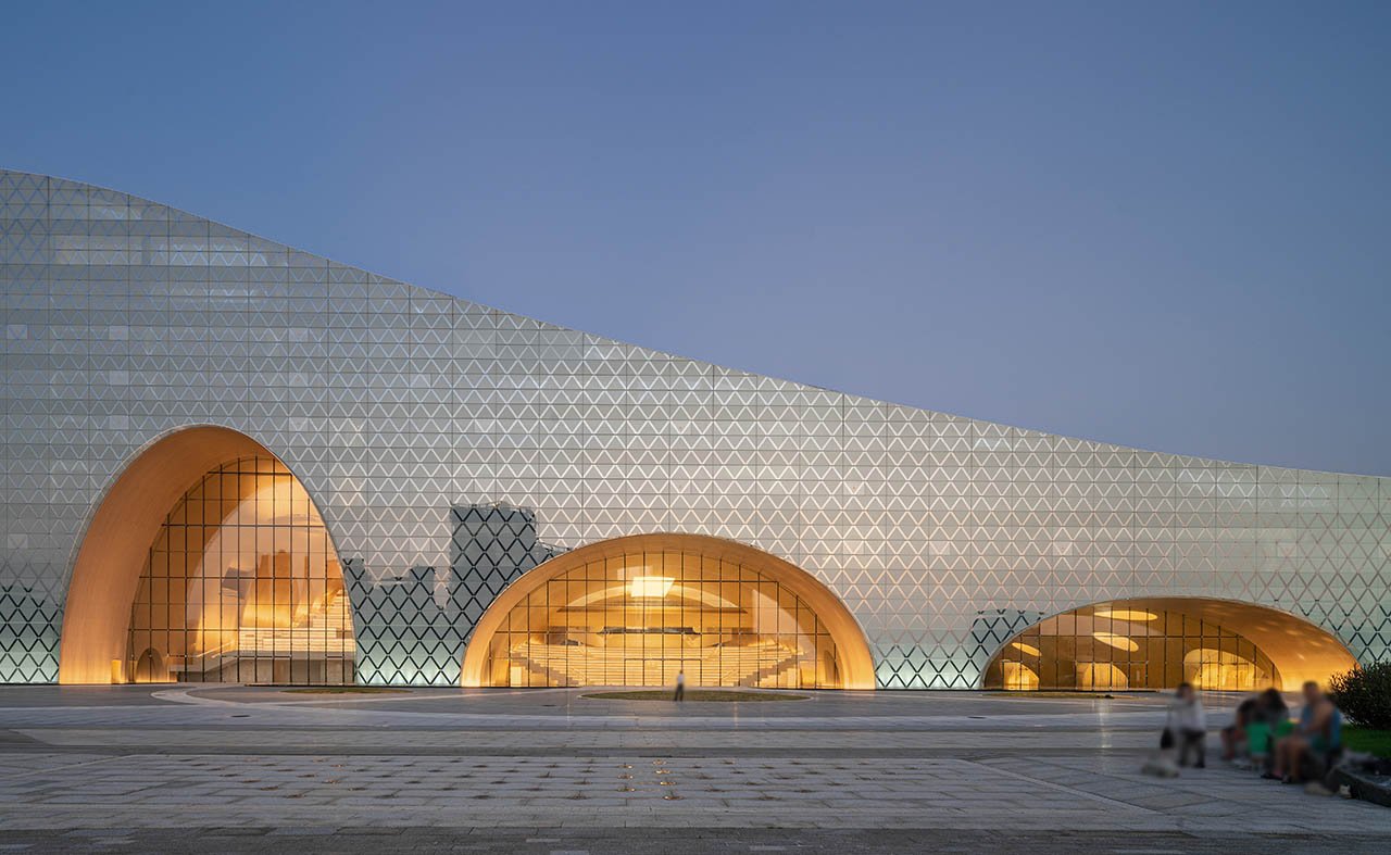 A modern architectural building with a unique design featuring large, curved archways and a facade covered in a geometric pattern of triangular shapes. The building emits a warm glow from within through its glass windows, creating a sleek and reflective contrast against the evening sky. A paved plaza is visible in the foreground, with a few people sitting and walking, which adds a sense of scale to the structure.