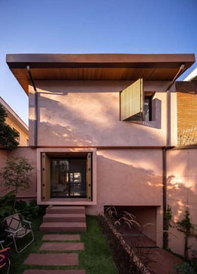 Modern two-story residence with a minimalist façade featuring light pink stucco and clean geometric lines. The upper level includes a large wood-slatted shutter over a wide window, while the ground floor showcases a recessed glass entrance flanked by wooden shutters and surrounded by a compact landscaped garden. A flat roof with wooden soffits and subtle overhang completes the contemporary aesthetic. Positioned between neighboring structures, the home contrasts warm textures with refined simplicity.