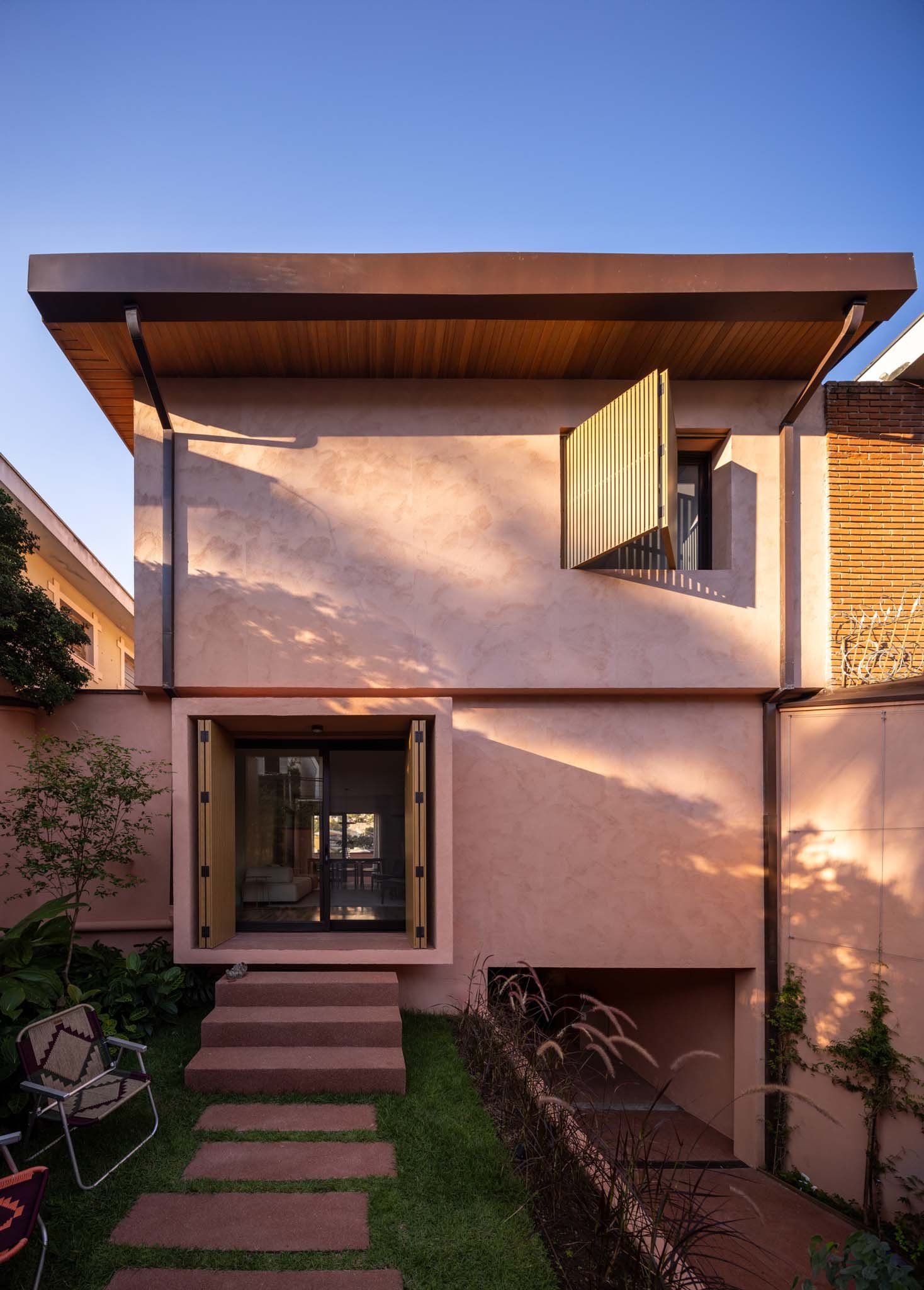 Modern two-story residence with a minimalist façade featuring light pink stucco and clean geometric lines. The upper level includes a large wood-slatted shutter over a wide window, while the ground floor showcases a recessed glass entrance flanked by wooden shutters and surrounded by a compact landscaped garden. A flat roof with wooden soffits and subtle overhang completes the contemporary aesthetic. Positioned between neighboring structures, the home contrasts warm textures with refined simplicity.