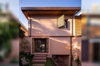 Modern two-story residence with a minimalist façade featuring light pink stucco and clean geometric lines. The upper level includes a large wood-slatted shutter over a wide window, while the ground floor showcases a recessed glass entrance flanked by wooden shutters and surrounded by a compact landscaped garden. A flat roof with wooden soffits and subtle overhang completes the contemporary aesthetic. Positioned between neighboring structures, the home contrasts warm textures with refined simplicity.