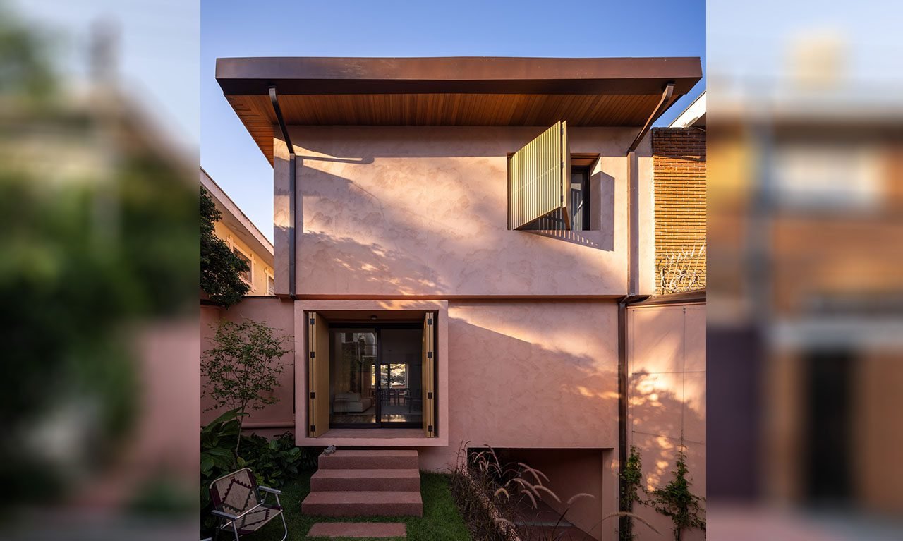 Modern two-story residence with a minimalist façade featuring light pink stucco and clean geometric lines. The upper level includes a large wood-slatted shutter over a wide window, while the ground floor showcases a recessed glass entrance flanked by wooden shutters and surrounded by a compact landscaped garden. A flat roof with wooden soffits and subtle overhang completes the contemporary aesthetic. Positioned between neighboring structures, the home contrasts warm textures with refined simplicity.